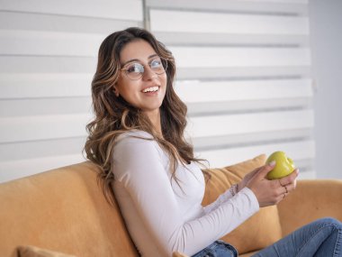 Sağlıklı beslenme konsepti, beyaz dişli, elinde taze yeşil elmayla gülümseyen beyaz kadın portresi evdeki kanepede oturmuş, sağlıklı beslenme konsepti için kamera arıyor. Boşluğu kopyala.