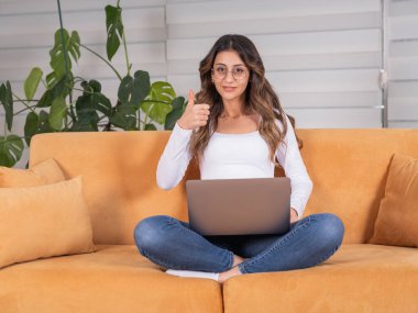 Baş parmak hareketi, tüm vücudu gören beyaz kadın kucağında dizüstü bilgisayarla kameraya gülümseyen bir jest sitesi göstererek oturma odasında kanepede oturuyor. Serbest kariyer kavramı.