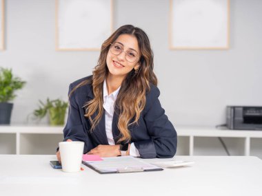 Mükemmel bir iş kadını, gülümseyen beyaz bir kızın portresi kameraya bakan kendine güvenen mükemmel bir iş kadını. Ofis masasında oturuyor, gözlük takıyor, resmi bir ceket giyiyor. Masada belgeler var..