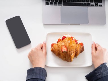 Lezzetli kruvasan, iş kadını ellerinin içinde lezzetli kruvasan olan tabağı tutuş görüntüsü. Siyah boş ekran cep telefonu maketi ve kesilmiş dizüstü bilgisayar. Ofis konseptinde kahvaltı.