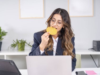 Kurabiye yiyen iş kadını, modern iç işyerinde dizüstü bilgisayar ekranı ararken 20 'li yaşlarda çekici beyaz bir iş kadını kurabiye yiyor. Boşluğu kopyala.