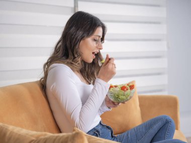 Evde salata yemek, evde salata yiyen 20 'li yaşlardaki genç beyaz bir kadının yan görüntüsü. Oturma odasında kanepede ya da kanepede bir kase organik sebze, vejetaryen atıştırmalığı ya da öğle yemeği bulundurmak. Boşluğu kopyala.