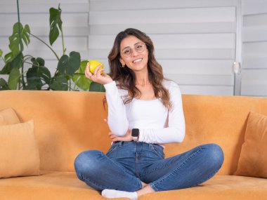 Vejetaryen bir kadın, tüm vücuduyla sağlıklı dişlerle gülümseyen taze yeşil elma tutan beyaz vejetaryen bir kadın. Oturma odasında kanepe ya da kanepe. Sağlıklı yaşam tarzı fikir görüntüsü.