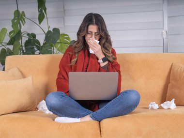 Sneezing woman, full body view young caucasian brunette sick ill sneezing woman sit at home couch or sofa using laptop wrapped in warm scarf. Sick and nauseous female working from house. clipart