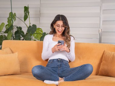 Heyecanlı mutlu kadın telefon kullanıyor, tüm vücudu görüyor... Kafkasyalı esmer, neşeli, mutlu kadın... evde oturma odasındaki koltukta oturuyor. Mesaj okunuyor, iyi haberler alınıyor.