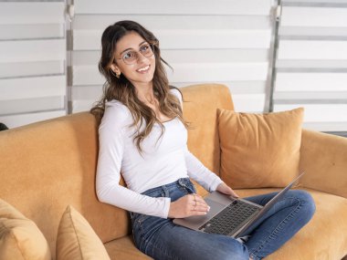 Kanepede oturan ve dizüstü bilgisayar kullanan bir kadın. Kafkasyalı, rahat giyinen, koltukta oturan ve internette dizüstü bilgisayar kullanan bir kadın. Genç yirmili yaşlardaki kızlar kanepede oturur..