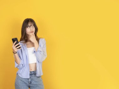 Genç düşünceli düşünceli kadın cep telefonunu sarı izole edilmiş arka planda duruyor çeneye dokunuyor çalışma alanı tanıtım alanına bakıyor, kopyalama alanına bakıyor. Halk yaşam tarzı konsepti.