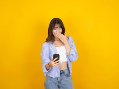 Şaşırmış genç esmer kadın cep telefonu kullanıyor, internetten mesaj yazıyor, sohbet ediyor, gülen yüz ifadesi takınıyor, beyaz bluz pantolon giyiyor, izole edilmiş sarı arka plan, kopyalama alanı kullanıyor.. 