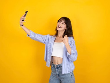 20 'li yaşlarda, mutlu, esmer, beyaz bir kadın izole edilmiş sarı stüdyo arka planında duruyor cep telefonuyla selfie çekiyor.. 