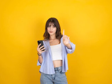 Akıllı telefon tutan güzel, çekici, beyaz, 20 'li yaşlarda, esmer, gülümseyen bir kadının portresi sarı stüdyo arka planında izole edilmiş bir kamerayı işaret ediyor. Boşluğu kopyala.