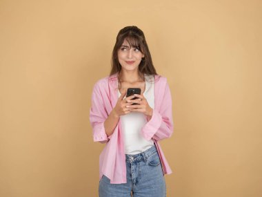 Tatlı genç bir kadın yana bakıyor, genç beyaz, tatlı bir kadının elinde cep telefonuyla gülümseyen bir yüz ifadesiyle arka plandaki izole edilmiş bej fotokopi alanının üzerinde durduğu fotoğrafı..