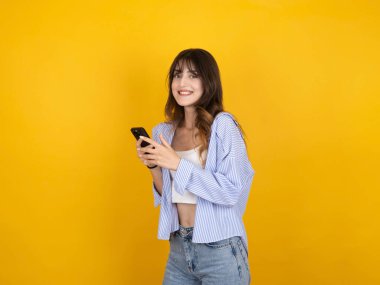 Telefon kullanan neşeli genç bir kadın, gülümseyen beyaz bir kadının portresi mutlu, genç bir kadın telefonla konuşuyor sarı stüdyo arka planında izole edilmiş internet mesajları, yaşam tarzı konsepti.