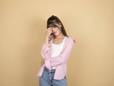 Dalgın, üzgün bir kadın, 20 'li yaşlarda, beyaz, esmer, uzun saçlı, dalgın, üzgün kadın pembe gömlek giyiyor, beyaz tişörtlü, gözlerini kapalı, burnu kapalı, bej arka planda, yaşam tarzı konseptinden soyutlanmış..