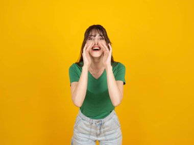 Çığlık atan bir kadın, 20 'li yaşlarda beyaz, esmer bir kadın, elleri ağızlarının yanında bağıran, izole edilmiş sarı arka plan stüdyo resmi. Heyecanlı mutlu yüz ifadesi insanların yaşam tarzı fikri.