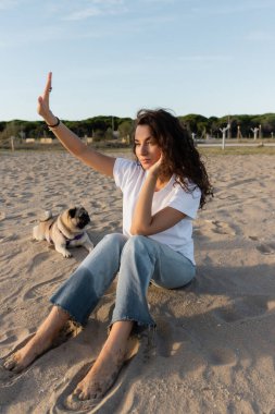 Kıvırcık, genç bir kadın, Barcelona 'da kumsalda, pug dog' un yanında uzanmış oturuyor. 