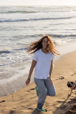 Gün batımında kumlu sahilde köpeğiyle koşan neşeli genç bir kadın. 
