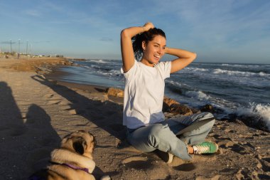 Neşeli serbest yazar dizüstü bilgisayarıyla oturuyor ve İspanya 'da deniz kıyısındaki köpek kumsalında kıvırcık saçlarını düzeltiyor. 