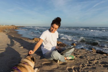 Telli kulaklıklı genç bir kadın dizüstü bilgisayarıyla oturmuş Barselona sahilinde köpeğe sarılıyor. 