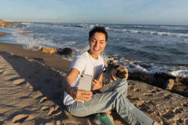 Telli kulaklıklı mutlu kadın dizüstü bilgisayar tutuyor ve Barcelona sahilinde köpeğiyle selfie çekiyor.
