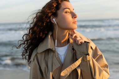 Trençkotlu kıvırcık kadın plajda telli kulaklıkla müzik dinliyor. 