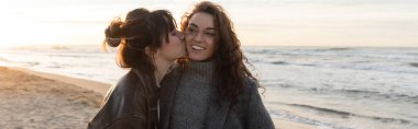 Young woman kissing cheeks of cheerful friend on beach in Spain, banner  clipart