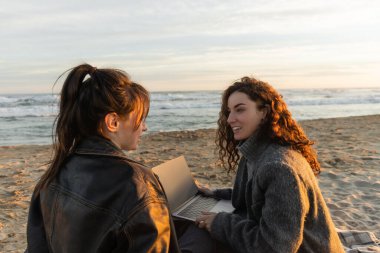 Smiling woman holding laptop while talking to friend on beach in evening  clipart