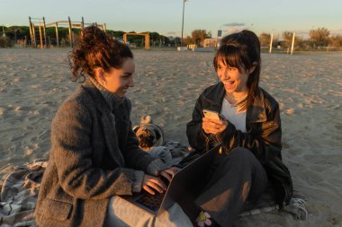 Cheerful women using gadgets near pug dog on beach in Spain  clipart