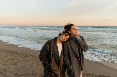 Young woman looking at camera near friend in coat on beach in Spain  clipart