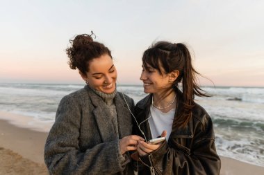 Gülümseyen arkadaşlar telli kulaklıkla müzik dinliyorlar ve kumsalda akıllı telefon tutuyorlar.  