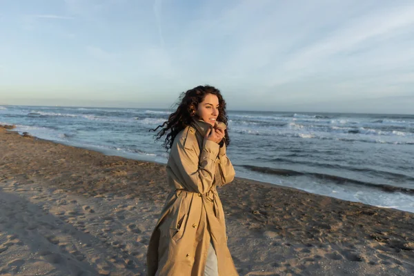 stock image cheerful woman in stylish beige trench coat walking on beach in Barcelona 