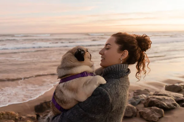 stock image Side view of smiling curly woman in coat holding pug dog on beach in Barcelona 