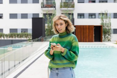 Barcelona 'da açık havuz otelinin yanında akıllı telefondan mesajlaşan kazaklı genç bir kadın.