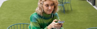 Yeşil kazaklı mutlu bir kadın telli kulaklıkla müzik dinliyor ve elinde akıllı telefon, afiş tutuyor. 