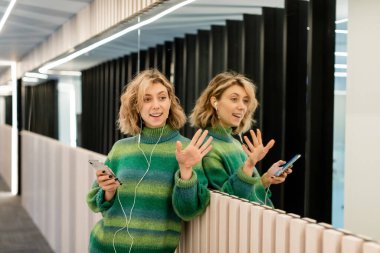 Telli kulaklıkla müzik dinleyen mutlu genç bir kadın oteldeki aynaların yanında el sallarken akıllı telefon tutuyor. 