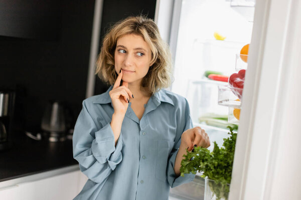 pensive young woman in blue shirt looking into open refrigerator in kitchen 