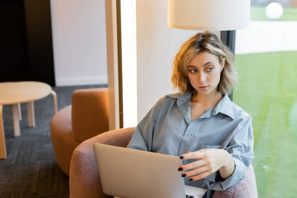 stock image pensive and blonde woman with wavy hair looking away while sitting in armchair with laptop 