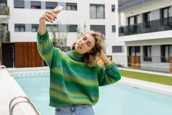 happy woman in sweater taking selfie on smartphone near outdoor swimming pool in Barcelona