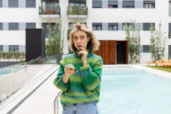 stock image pensive young woman in sweater holding smartphone near outdoor swimming pool of hotel in Barcelona