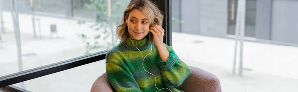 stock image cheerful woman in green sweater adjusting earphones while sitting in lobby of hotel in Barcelona, banner 