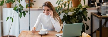 Vienna 'daki kafede bilgisayarın yanında akıllı telefondan konuşan neşeli bir kadın, afiş. 