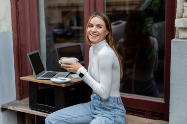 joyful freelancer holding cup of coffee near laptop with blank screen in cafe while sitting outdoors in Vienna  clipart