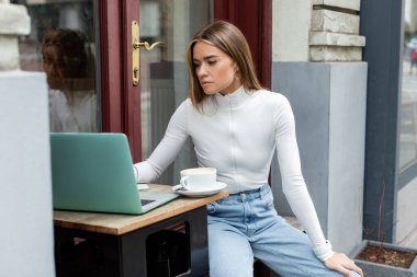 Viyana 'da açık bir kafede otururken bir fincan kahvenin yanında dizüstü bilgisayar kullanan genç bir kadın. 