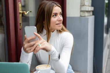 Gülümseyen genç bir kadın Viyana 'da kafede otururken bir bardak kapuçino yanında akıllı telefon tutuyor. 