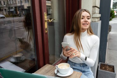 Gülümseyen genç bir kadın Viyana 'da açık bir kafede otururken bir fincan kahvenin yanında akıllı telefon tutuyor. 