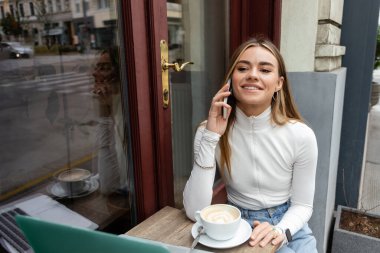 pleased woman talking on smartphone near cup of coffee while sitting in outdoor cafe in Vienna  clipart