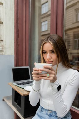 Viyana 'daki boş ekranlı laptopun yanındaki kafede oturan genç bir kadın kahve içiyor. 