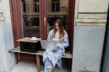 young freelancer in blue trench coat using laptop while sitting next to cup of coffee in outdoor cafe in Vienna   clipart