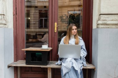 young woman in blue trench coat using laptop while sitting next to cup of coffee in outdoor cafe in Vienna   clipart