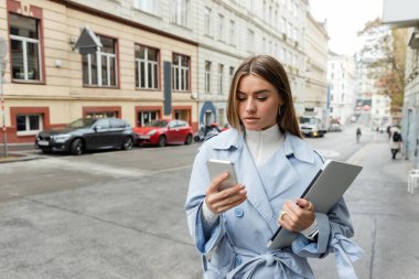 Mavi trençkotlu güzel bir kadın Viyana 'da dizüstü bilgisayar tutarken akıllı telefon kullanıyor. 