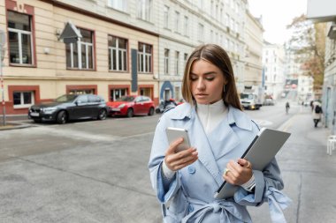 pretty young woman in blue trench coat using smartphone while holding laptop on street in Vienna  clipart